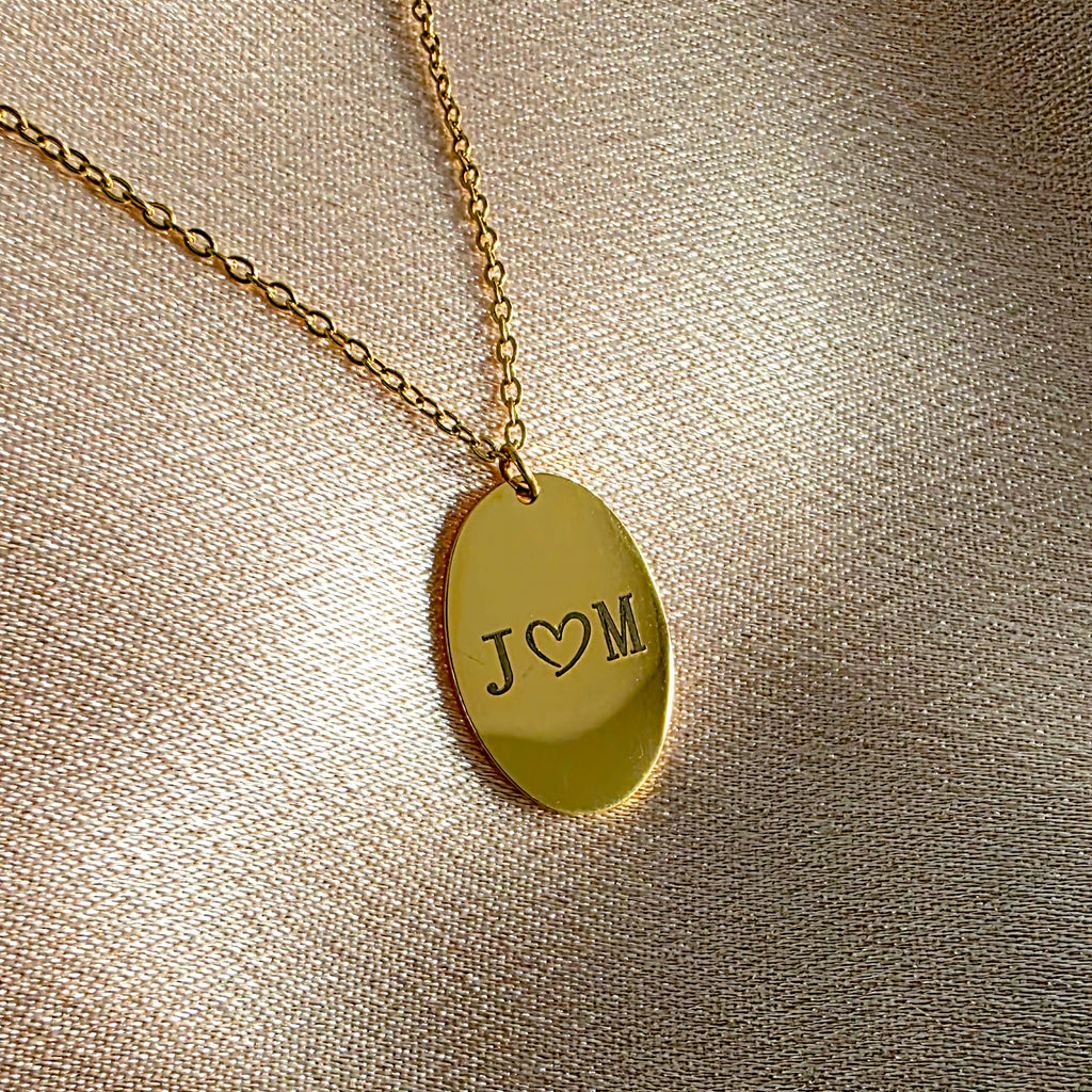 Gold necklace with a pendant engraved with 'J ❤️ M' on a beige fabric background
