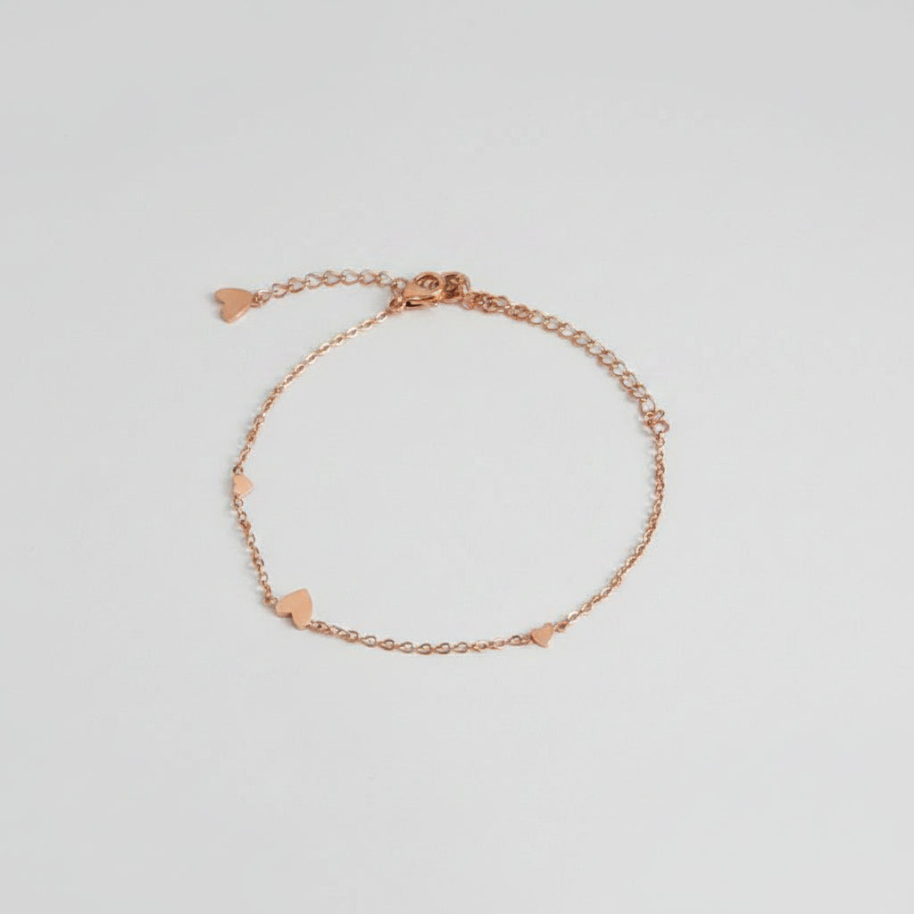 A delicate rose gold chain bracelet with small heart charms, displayed on a white surface next to a black pouch with white text and a clear glass bowl.