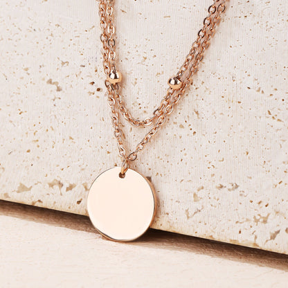 A rose gold double-layer necklace with a flat round disc pendant, displayed on a textured beige stone surface.