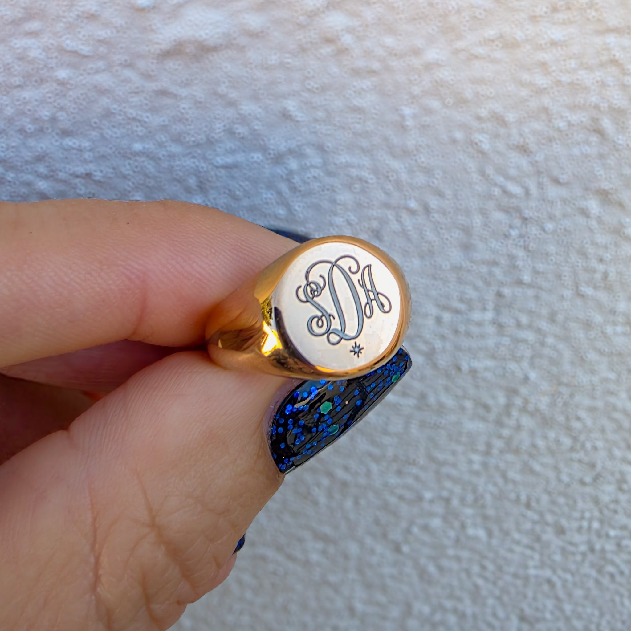 Rose gold signet ring with monogram design held by a hand against a textured beige background